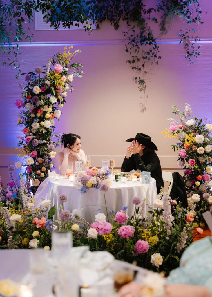 Bride and groom seated at a floral-filled sweetheart table featuring colorful garden-inspired blooms and hanging greenery at an elegant wedding reception.