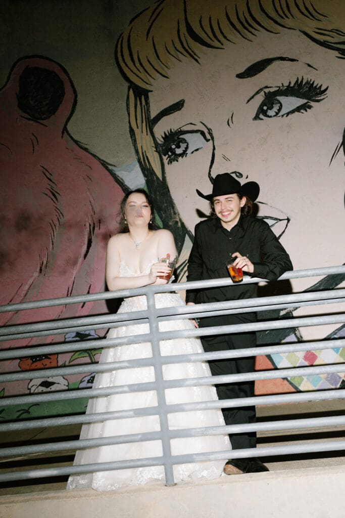 Newlywed couple poses with drinks in front of a historic downtown Brenham mural during nighttime wedding portraits at Ant Street Inn.