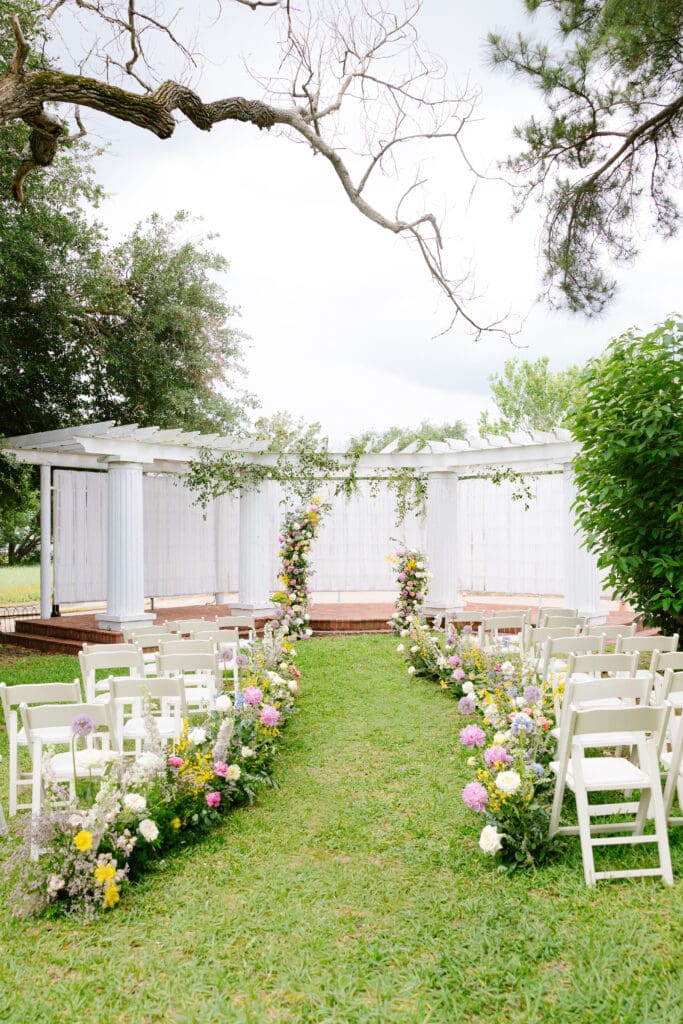 Garden wedding ceremony design at Ant Street Inn in Brenham, Texas with floral aisle arrangements and a white pergola backdrop.